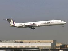 A jet airliner with tail-mounted engines and a T-tail configuration, painted mostly white with a thin red stripe running along the fuselage below the windows, flying low above airport buildings with landing gear and flaps extended.