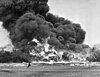 A demonstration of flame fougasse, somewhere in Britain. A car is surrounded in flames and a huge cloud of smoke. In the foreground, a soldier can be seen running from left to right.