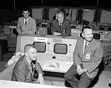 Photographie en noir et blanc de Kraft avec trois autres directeurs de vol près de leurs consoles.