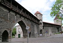 Seitliche Farbfotografie von einer alten, grauen Steinmauer, durch deren Rundbogen im Vordergrund eine Römerstraße führt. Auf der Mauer ist ein gedeckter Holzbau, der im rechten Hintergrund von zwei Wachtürmen unterbrochen wird.