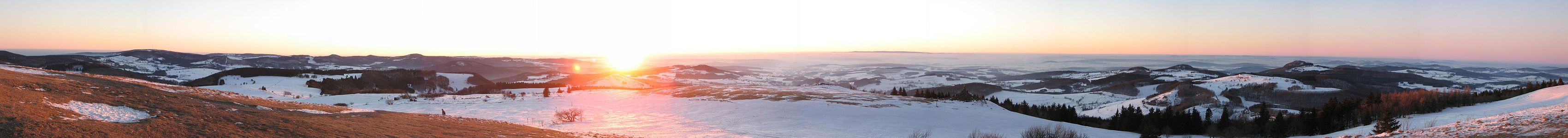 Sonnenuntergang unterhalb des Radoms. Hier sind Tische und Bänke vorhanden, von denen aus man den Westblick mit Sonnenuntergang genießen kann. Zum Panorama: Aufgrund einer Inversionswetterlage herrscht sehr gute Fernsicht. Der Berg mit Funkturm ganz links im Bild ist der Kreuzberg. Rechts neben der Sonne liegt am Horizont der Vogelsberg (Entfernung etwa 45 km). Zwischen Sonne und Vogelsberg erkennt man in der Bildervergrößerung übrigens sogar den Grossen Feldberg im Taunus (Luftlinie etwa 110 km). Rechts im Vordergrund liegt die Kuppenrhön mit der markanten Erhebung der Milseburg. Noch weiter Rechts ist am Horizont ein länglicher Höhenzug zu erkennen. Dies ist der Hohe Meißner (Entfernung etwa 80 km).