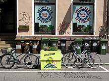 Zwei Fahrräder stehen beidseitig in Fahrradständern eines bunt bemalten Hochbeetes, das auf der Markierung eines Pkw-Stellplätzes steht. Im Hintergrund die Fassade eines Ladengeschäfts