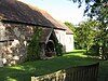 Part of the south face of a small stone church, seen from an angle, with tiled roofs and a gabled, wooden porch