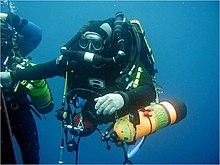 A scuba diver in a wetsuit holds onto the shotline at a decompression stop. He is breathing from a rebreather and carrying a side-slung 80 cubic foot aluminium bailout cylinder on each side. A second diver is partly visible to the left.