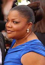 An African American female with dark brown hair up in a bun with a flower. She is wearing a short sleeved royal blue dress.