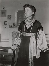 ca. 1903 aufgenommenes Foto, auf dem Gabriele Münter mit ernstem Blick in einem Wohnzimmer steht