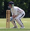 A woman wearing white cricket uniform with red piping and a blue helmet crouches behind three wooden stumps.