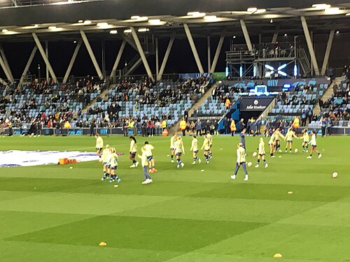 Manchester City W.F.C. players wearing yellow pre-match jerseys, with small details of blue, warm up in front of the Joie Stadium's blue stands