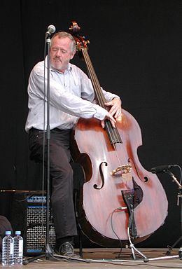 Le contrebassiste Niels-Henning Ørsted Pedersen est vêtu d'un pantalon noir et d'une chemise sombre. Il est sur scène, assis sur un taouret haut, en train de jouer de la contrebasse. Les yeux fermés, la tête tournée vers sa droite, ses mains sont dans le registre aigu de son instrument.