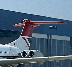 Photographie en couleurs d'une aile, d'un moteur et de la dérive rouge d'un avion, devant un hangar.