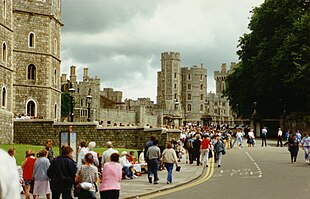 A photograph of a grey stone castle, stretching alongside the left of the picture. A road makes up the centre, along which a number of people walk.