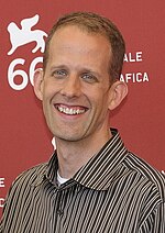 A blonde man is smiling. He wears a stripped shirt, and he is standing in front of a red wall.