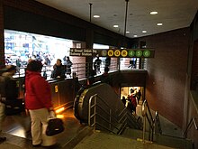 The subway entrance at the ground story of the Zeckendorf Towers. There are escalators to the left and stairs to the right. A sign with the station's name, and the services that stop at the station, is placed above the escalators and stairs.