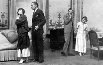 Stage scene, with middle-aged white man and women to left, in discussion, and two younger adults tiptoeing away