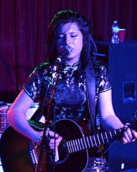 Chloe Chaidez performing on stage; singing into a microphone and playing a guitar