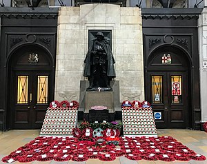 Bronze statue against a white stone wall with oak panels in the background and poppy wreaths on the floor below