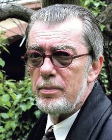 Publicity shot of elderly man in three-quarters profile, with short, trimmed white beard and swept-back, short grey hair. He is wearing dark glasses.