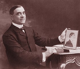 Middle aged white man, clean shaven, in smart suit sitting at a desk holding a caricature of himself