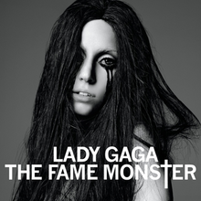 A woman in a blonde blunt bob cut. She wears a shiny black leather dress covering her body completely. With her right hand she holds the collar of the dress and uses it to cover her mouth. In front of her, the words "Lady Gaga" and "The Fame Monster" are written in white capital font, with the T of Monster being stylized as a cross (†).