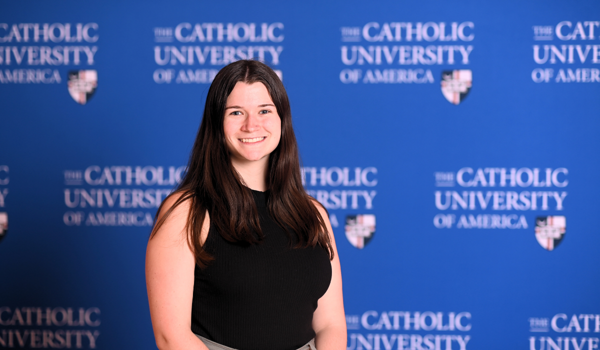 Maria Duffy is standing infront of a blue Catholic University background. She has long dark hair and is wearing a black sweater.