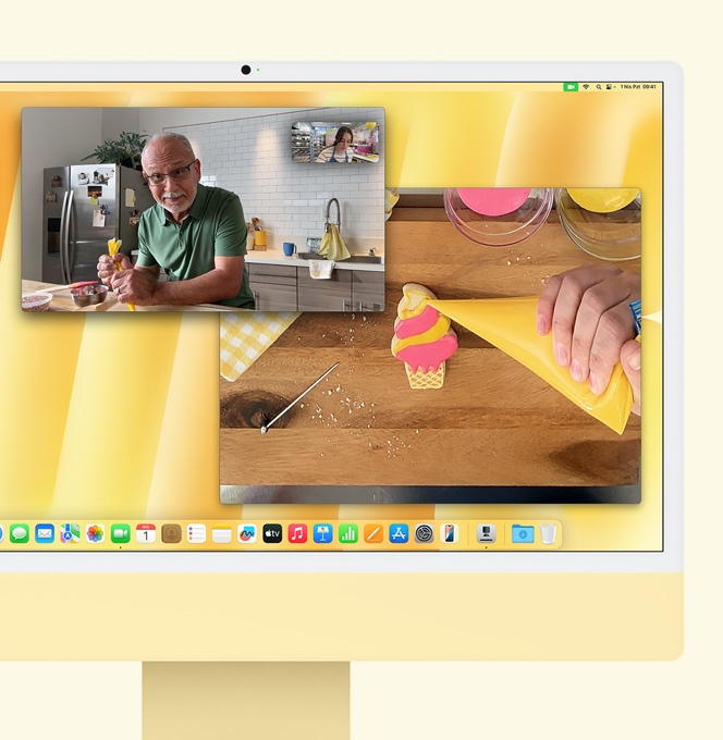 Yellow iMac. Desk View demo. Screen shows baker on video call with another person. One screen displays the baker and the man's face. The other screen displays the baker's hands icing a cookie