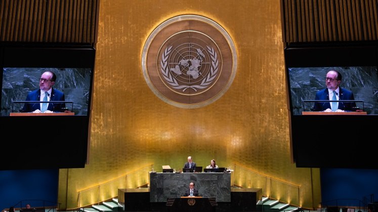 The picture shows Austria's Foreign Minister Alexander Schallenberg at the lectern in front of the UN General Assembly. The logo of the United Nations can be seen on the wall above Foreign Minister Schallenberg. A close-up of Schallenberg is shown on a screen to the left and right of the logo.