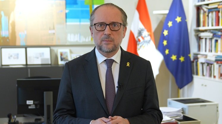 Außenminister Schallenberg steht in seinem Büro und blickt in die Kamera. Im Hintergrund sind die Flaggen der Europäischen Union und Österreichs zu sehen.