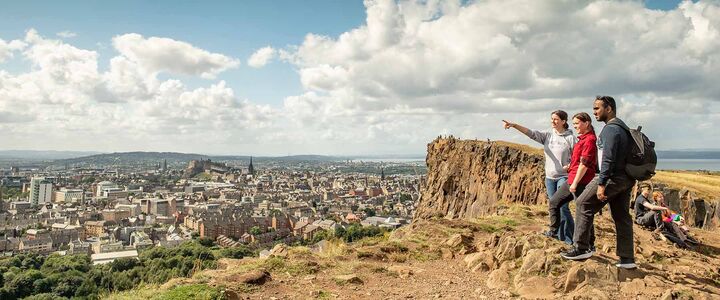 Three students stand on top of Salisbury Crags overlooking Edinburgh city