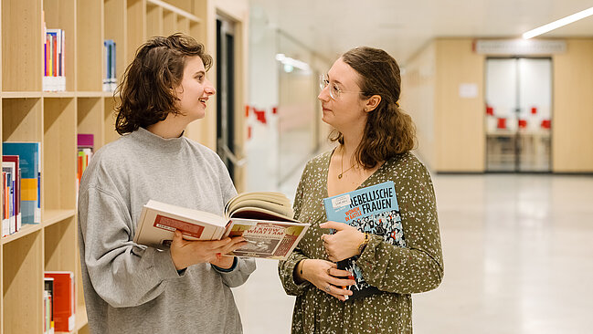 Foto von zwei Personen, die sich unterhalten und dabei Bücher halten.