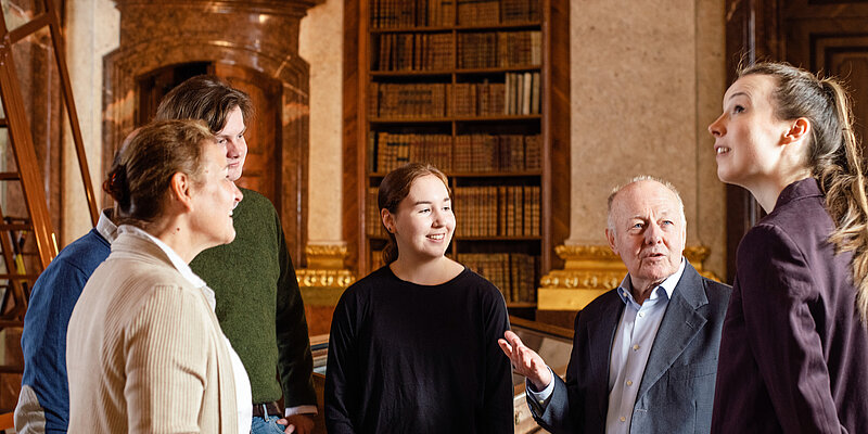 Eine Gruppe von Menschen nimmt an einer Führung in einem barocken Bibliothekssaal teil.