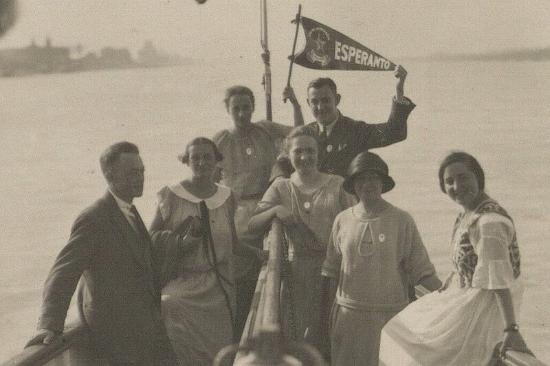 Altes, schwarz-weißes Foto von lächelnden Personen am Bug eines Schiffs am Wasser, eine Person hält eine Esperanto-Flagge.