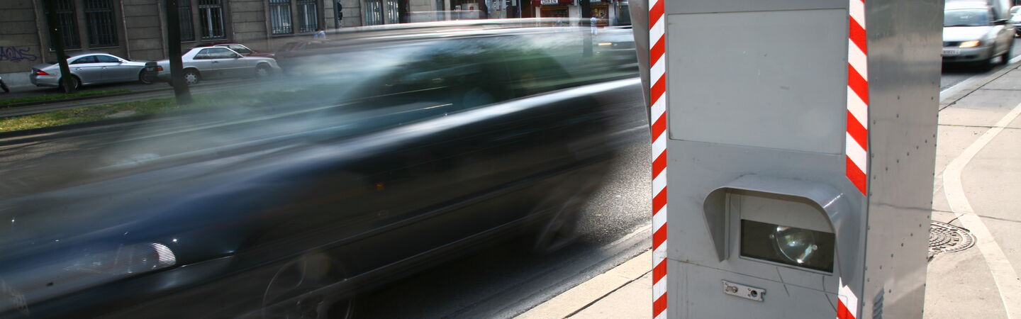 Rechts im Bild sieht man eine Radarbox, bei der ein schwarzes Auto auf der Straße vorbeifährt.