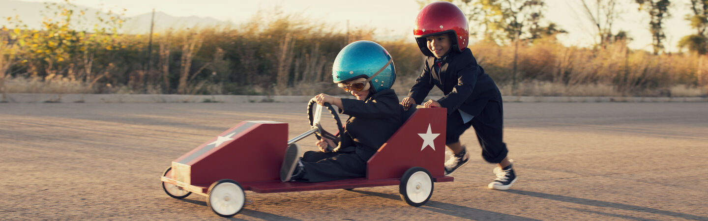 Zwei Kinder mit großen Helmen haben Spaß mit einem lustigen Raketenfahrzeug. Ein Kind mit blauem Helm sitzt im Fahrzeug, das zweite Kind mit rotem Helm schiebt hinten das Gefährt an.