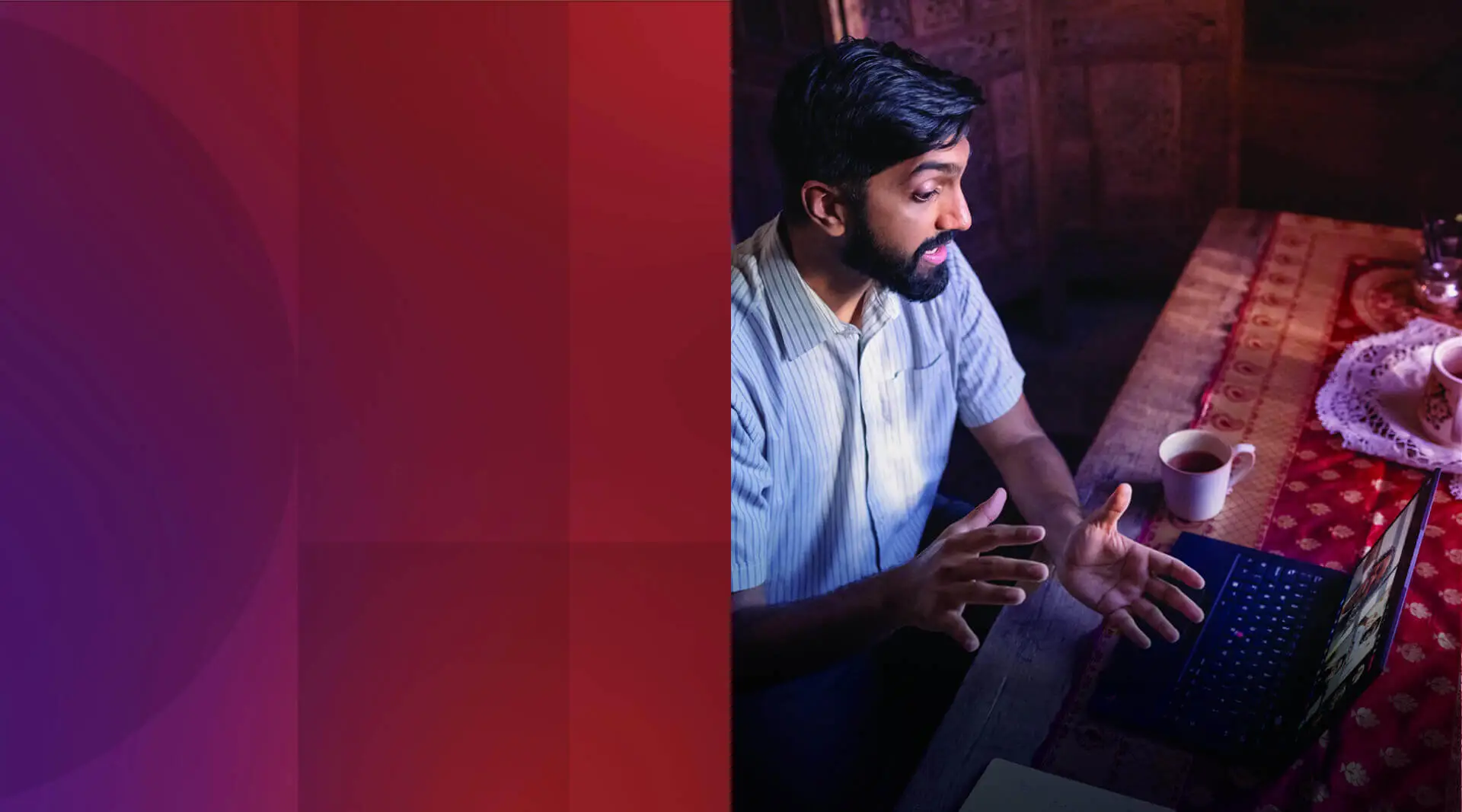 A bearded, dark-haired person in a light blue shirt makes hand gestures in a restaurant booth as he talks during a video call on his laptop. 