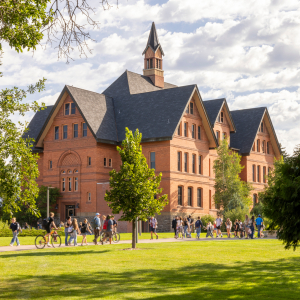 The image shows a large group of students walking and biking across the green, tree-lined lawn of Montana State University. In the background is the iconic brick structure of Montana Hall, with its steep gabled roofs and central cupola. The sky is partly 