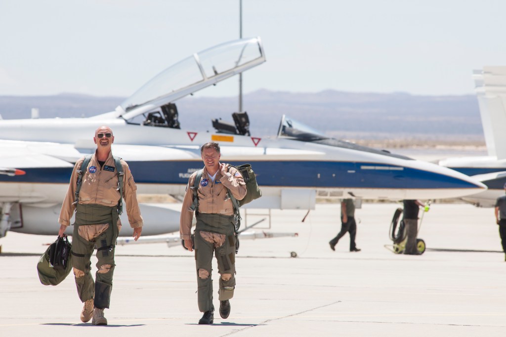 Two NASA test pilots walk toward the camera. Behind them is a blue and white F/A-18 research jet.