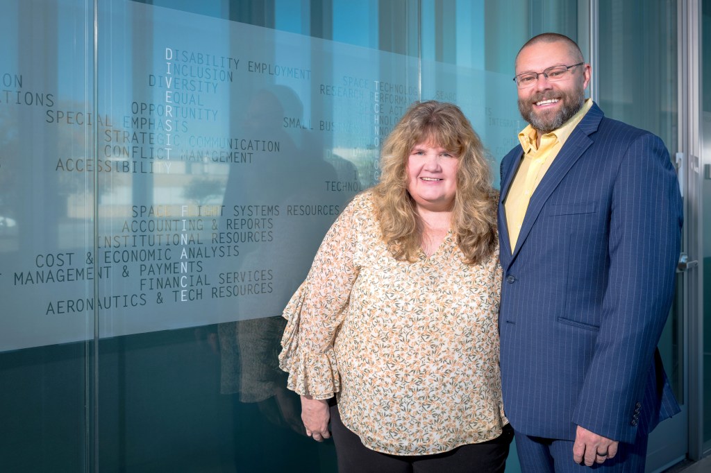 A man wearing a blue suit, a yellow shirt, and glasses and a woman wearing a yellow blouse smile at the camera as they pose standing side by side in front of a glazed glass wall. Small words are etched on the wall, including “Disability Employment, Inclusion, Diversity, Equal Opportunity, Accessibility,” and more.