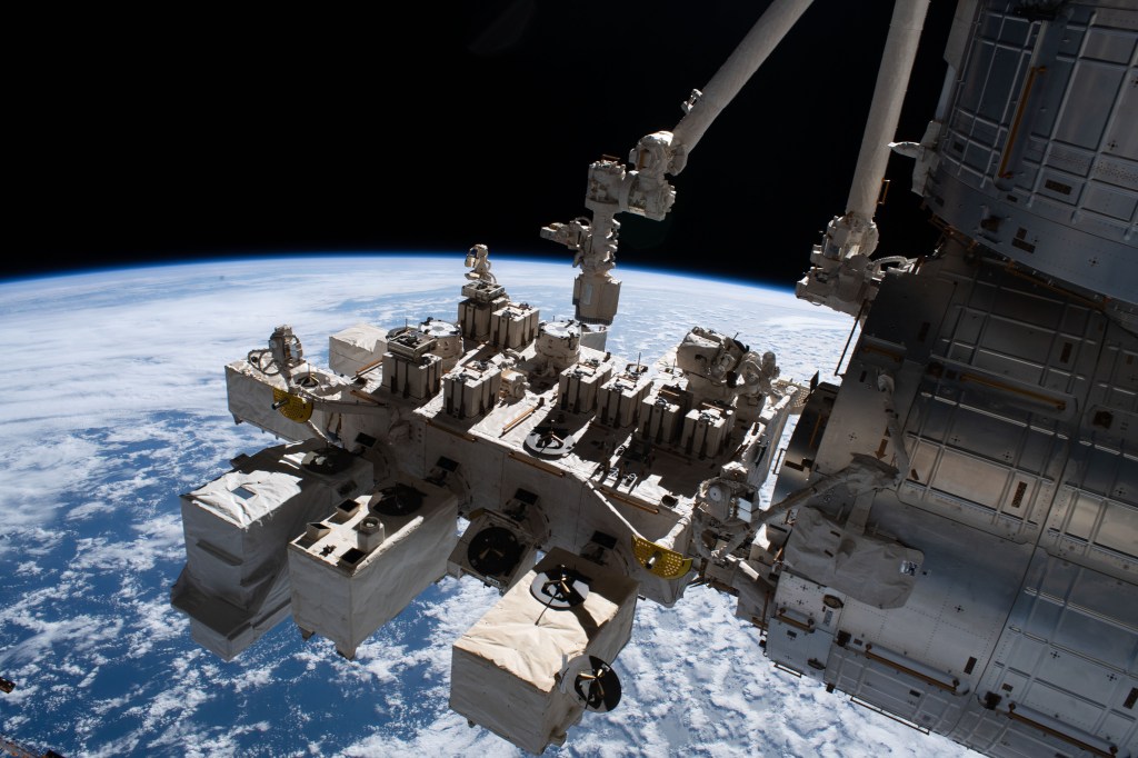 Exterior view of the International Space Station with Earth visible in the background. The station’s robotic arm and multiple scientific equipment modules are attached to its surface.