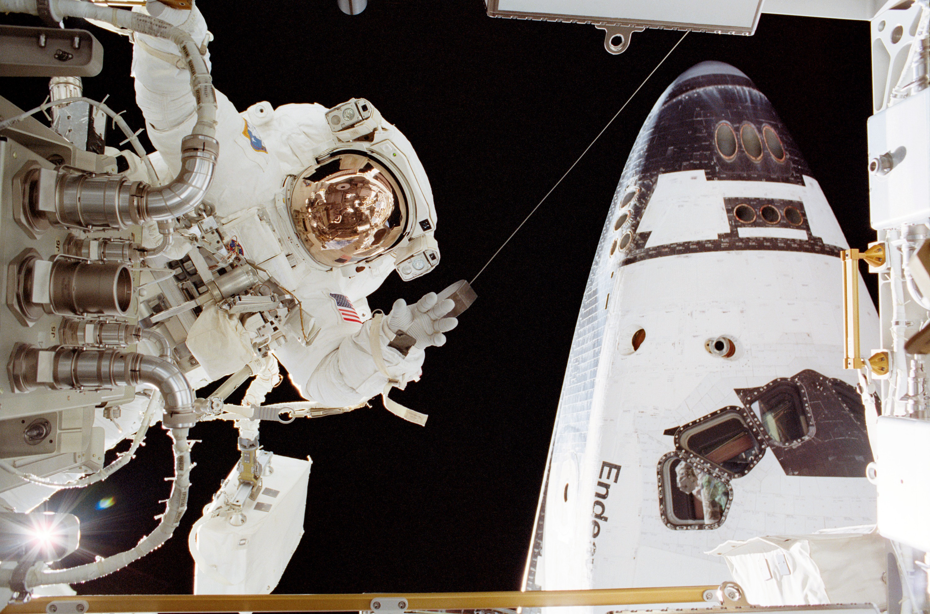 An astronaut in a white spacesuit appears on the left side of the image near a part. On the right in the back is part of the space shuttle Endeavour.