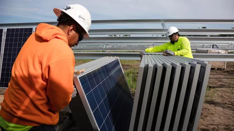 Two workers wearing hard hats separating a stack of solar panels at a construction site