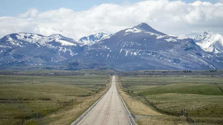 A road surrounded by vast fields of grass on either side, with snowcapped mountains in the distance.