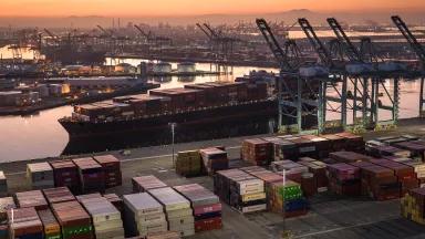 An aerial view of stacked shipping containers and a full container ship in the Port of Los Angeles at sunrise