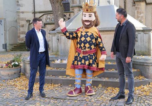 Zwei Männer und in ihrer Mitte ein Königsmaskottchen vor Brunnen