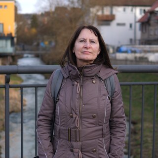 Frau mit braunen Haaren steht auf Brücke, unter der ein kleiner Bach fließt. Herbstliche Anmutung.