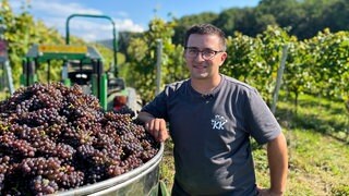 Christopher ist Jungwinzer im Nebenerwerb. Er steht neben einem Tank, der mit Graburgunder gefüllt ist. Er lacht in die Kamera.