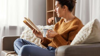 Eine Frau sitzt im Schlafanzug auf einer Couch und liest ein Buch. In der Hand hält sie eine Tasse Kaffee.