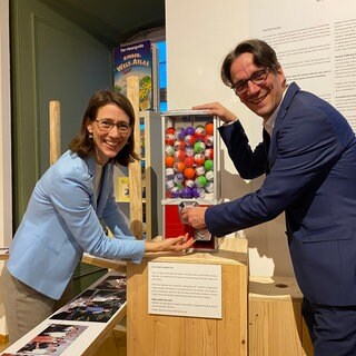 Bettina Gräfin Bernadotte und ihr Bruder Björn Graf Bernadotte stehen an einer Art Kaugummi-Automaten, in dem sich Kugeln mit Zetteln befinden. Auf den Zetteln stehen Witze. Eine Kindheitserinnerung der beiden.