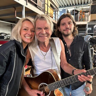 Matthias Reim steht mit Gitarre um den Hals zwischen Ehefrau Christin Stark und Sohn Julian Reim. Sie lachen in die Kamera.