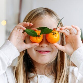 Ein blondes Mädchen hält sich lächelnd zwei Mandarinen vor die Augen: Eine gesunde, vitaminreiche Ernährung ist wichtig für die Augen