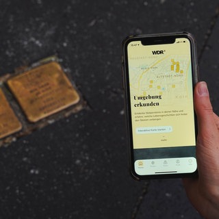 Eine Hand mit Smartphone, auf dem die App "Stolpersteine" zu sehen ist